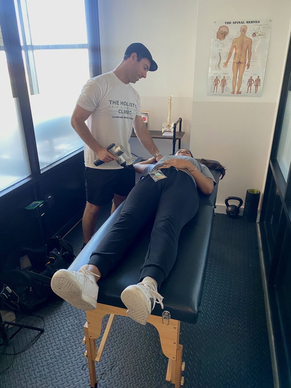 Hands-on bodywork with a massage gun in a movement-focused treatment space — table care alongside rehab tools
